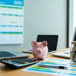 A desk with a laptop, calculator, and colorful budgeting spreadsheets. A piggy bank and a jar of coins sit nearby. A calendar on the wall shows the year 2023